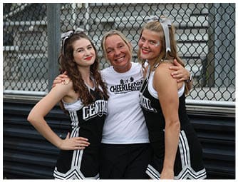 Three people smiling and posing together with two wearing cheerleading uniforms and one wearing a cheerleading coach shirt in front of a chain-link fence and bleachers. Descripci n generada con IA