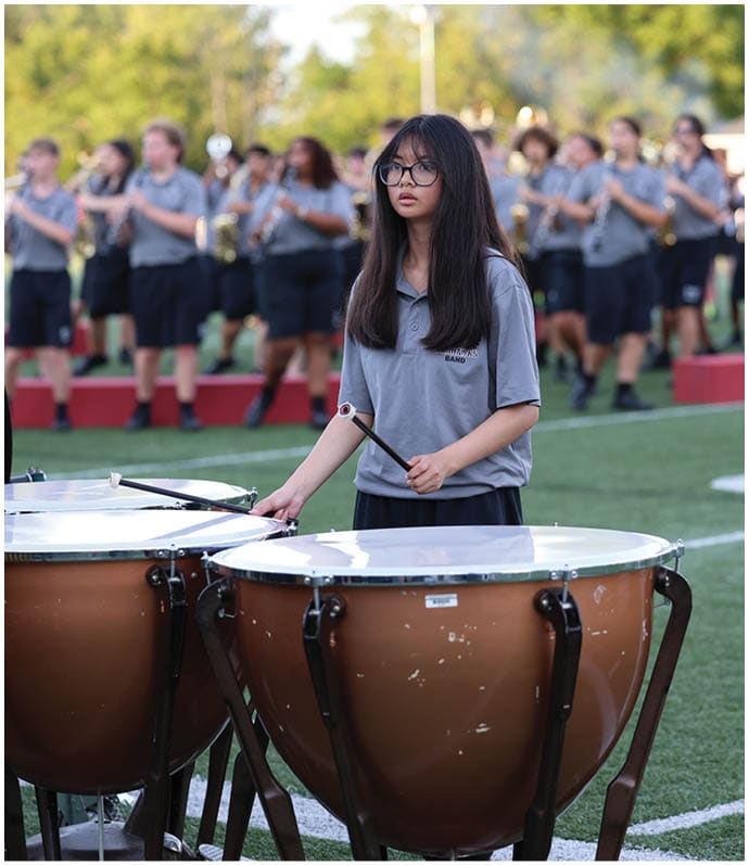 A person with long hair and glasses wearing a gray polo shirt is playing a timpani drum on a sports field with a marching band performing in the background. Descripci n generada con IA