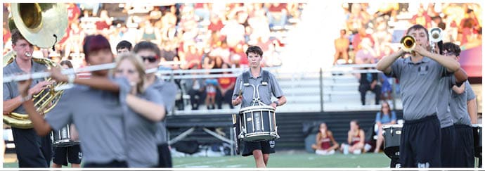 People in a marching band uniform are playing instruments on a field with a crowd in the background. Descripci n generada con IA