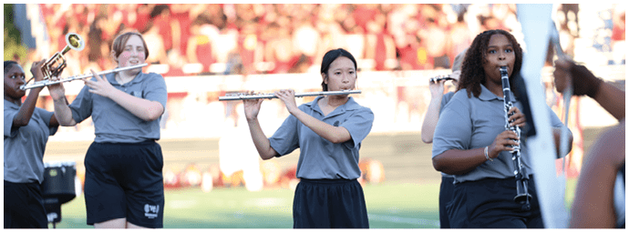 People wearing gray shirts and black pants are playing wind instruments in an outdoor setting with a blurred crowd in the background. Descripci n generada con IA