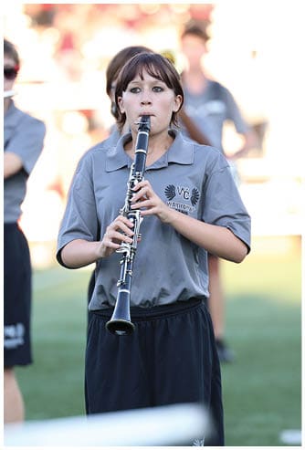 A person wearing a gray polo shirt and black shorts is playing a clarinet on a grassy field. Descripci n generada con IA