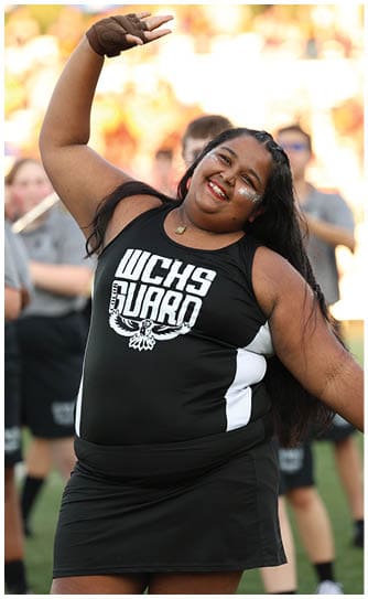 A person with long hair wearing a black and white cheerleading uniform with “UCHS Guard" printed on the front is smiling and posing outdoors. Descripci n generada con IA