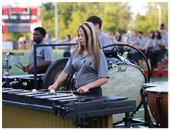 People wearing matching gray shirts and black shorts are playing flutes outdoors on a field with a blurred crowd in the background. Descripci n generada con IA