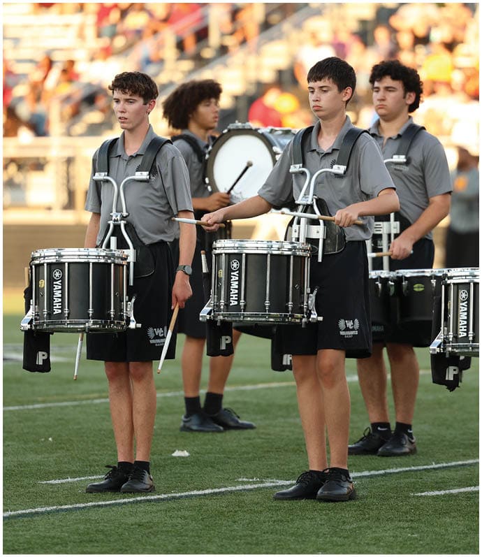 Four people wearing matching gray shirts and black shorts are playing drums on a field with a blurred crowd in the background. Descripci n generada con IA