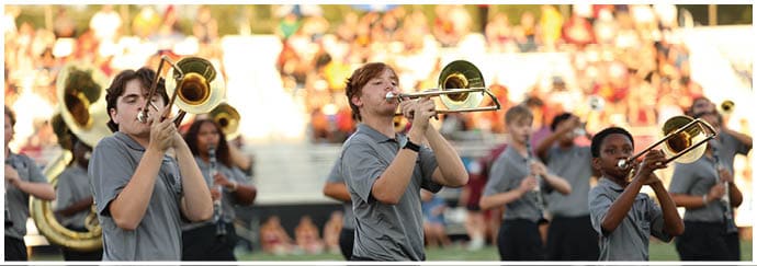 People wearing gray shirts are playing trombones outdoors on a field with a crowd in the background. Descripci n generada con IA