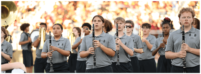 A group of people wearing matching gray shirts are standing outdoors playing clarinets in a band setting with a blurred crowd in the background. Descripci n generada con IA