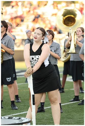 A person in a black and white cheerleading uniform stands on a sports field holding a white baton with a marching band playing instruments in the background. Descripci n generada con IA