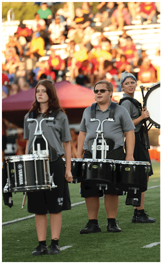Two people wearing gray shirts and black pants are standing on a sports field playing percussion instruments with a blurred crowd in the background. Descripci n generada con IA