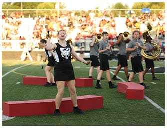 A person in a black and white uniform stands on a red platform on a sports field holding a baton with a crowd and other band members playing instruments in the background. Descripci n generada con IA