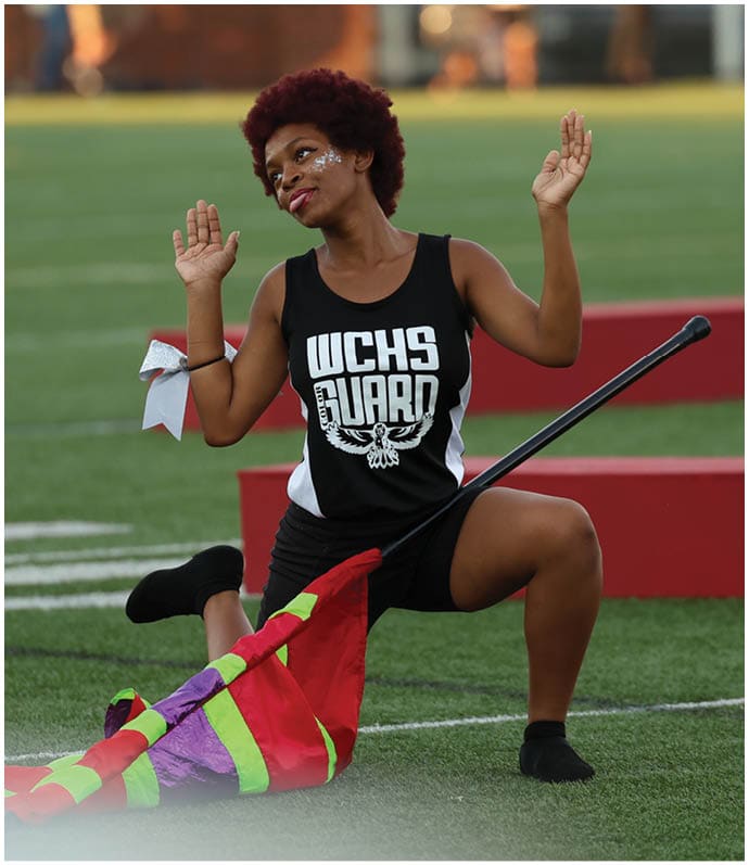 A person with short curly hair wearing a black tank top with “WCHS Guard" printed on it is kneeling on a sports field holding a colorful flag. Descripci n generada con IA