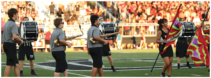 People in a marching band are playing drums and saxophones while others carry colorful flags on a sports field with a crowd in the background. Descripci n generada con IA