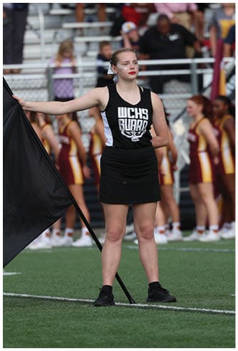 A person wearing a black WCHS Guard uniform stands on a sports field holding a black flag with a group of people in maroon and yellow uniforms blurred in the background. Descripci n generada con IA