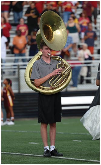 A person wearing a gray shirt and black shorts is playing a sousaphone on a sports field with a crowd in the background. Descripci n generada con IA