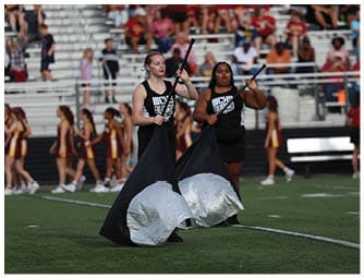 Two adults in black uniforms holding flags perform on a sports field with a crowd and cheerleaders in the background. Descripci n generada con IA
