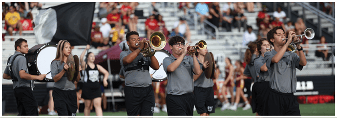 People in matching gray shirts are playing brass and percussion instruments in a marching band with a crowd in the background. Descripci n generada con IA