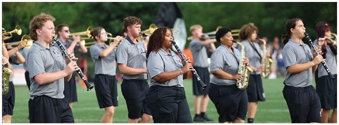 A group of people wearing matching gray shirts and black shorts are playing clarinets and saxophones outdoors on a grassy field. Descripci n generada con IA