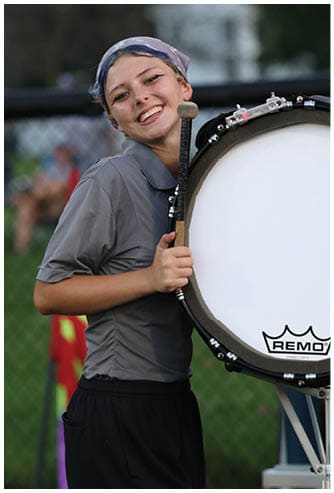 A person wearing a headscarf and casual clothing is smiling while holding a large bass drum and a drum mallet outdoors near a fence. Descripci n generada con IA
