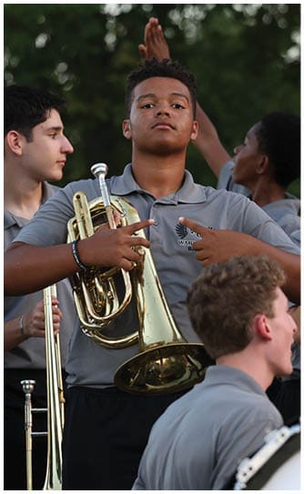 A group of people wearing matching gray shirts are gathered outdoors with musical instruments including a trumpet, trombone, and drum, appearing to prepare for a band performance. Descripci n generada con IA