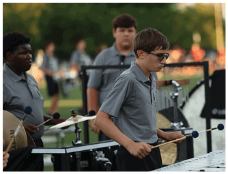People wearing matching gray shirts are playing percussion instruments outdoors, with one person focused on a xylophone and others playing cymbals and drums. Descripci n generada con IA