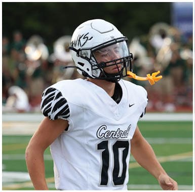 A person wearing a white football uniform with the number 10 and a helmet is standing on a football field with a band playing in the background. Descripci n generada con IA