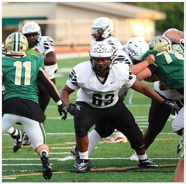 People in white and green football uniforms are engaged in a play on a football field with a blurred background of trees and a building. Descripci n generada con IA