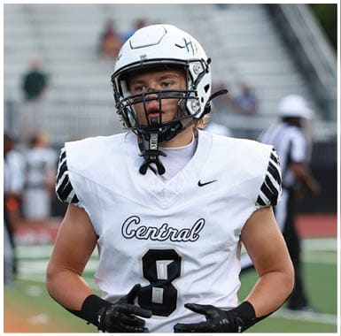 A person wearing a white football uniform with the word Central and the number 8 on the jersey is standing on a football field with blurred figures and bleachers in the background. Descripci n generada con IA