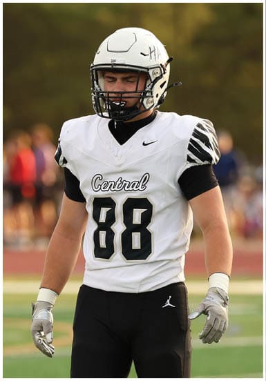 A person wearing a white football helmet and a white and black football uniform with the number 88 stands on a green football field with blurred spectators and a referee in the background. Descripci n generada con IA