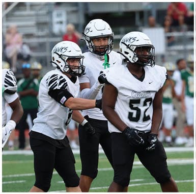 Three people in white football uniforms and helmets stand on a field with blurred spectators in the background. Descripci n generada con IA