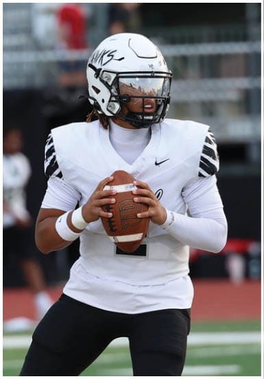 A person wearing a white football uniform and helmet is holding a football on a field with blurred spectators and players in the background. Descripci n generada con IA