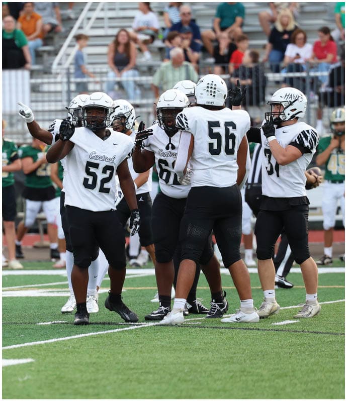 A group of football players in white and black uniforms are gathered on a field with spectators in the background. Descripci n generada con IA