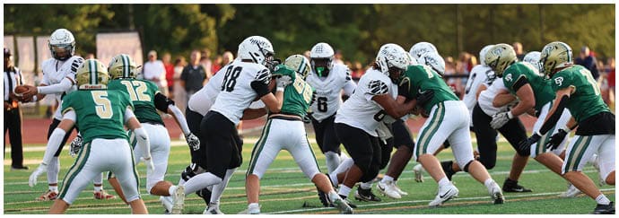 People wearing football helmets and uniforms are engaged in a play on a grassy field with spectators in the background. Descripci n generada con IA