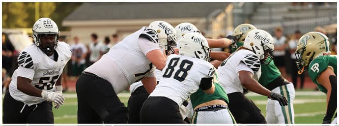 People wearing football uniforms are engaged in a play on a football field with some players blocking and others preparing to move. Descripci n generada con IA
