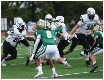 Several people wearing football uniforms are actively engaged in a play on a green field with a blurred background of spectators and trees. Descripci n generada con IA