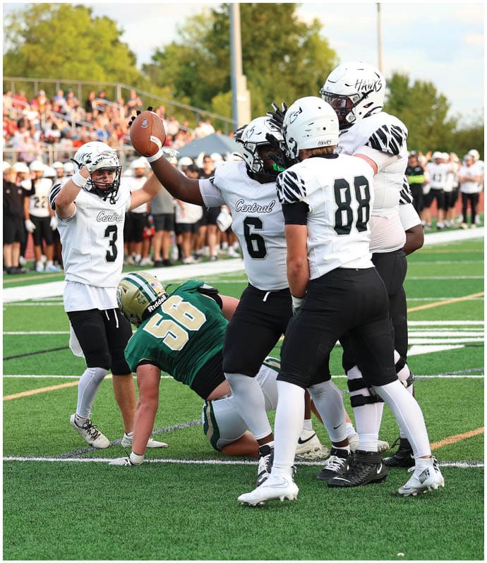 A group of football players in white uniforms celebrate on a field while a player in a green uniform kneels on the ground during a game with a crowd in the background. Descripci n generada con IA