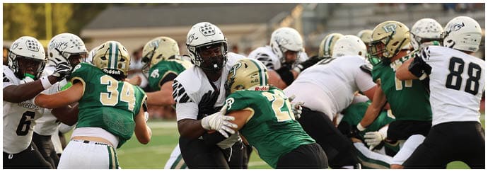 People wearing football uniforms are engaged in a play on a grassy field with some players blocking and others preparing to move. Descripci n generada con IA