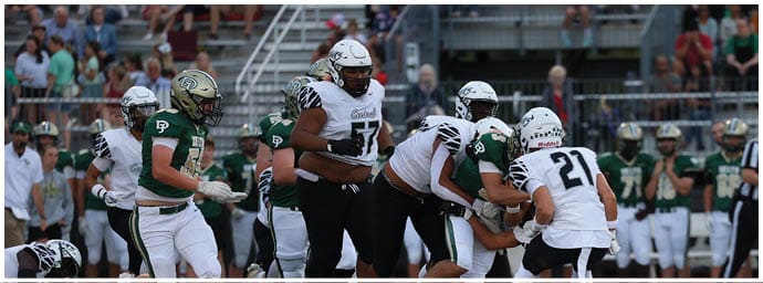 People wearing football uniforms are engaged in a play on a football field with some players tackling and others running. Descripci n generada con IA