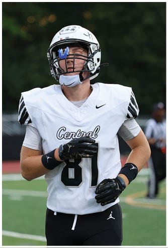 A person wearing a white sports jersey with black gloves and a wristband is on a football field with a blurred referee in the background. Descripci n generada con IA