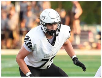 A person in a white football uniform with a helmet and knee brace is positioned on a football field facing two opponents in green uniforms during a game. Descripci n generada con IA