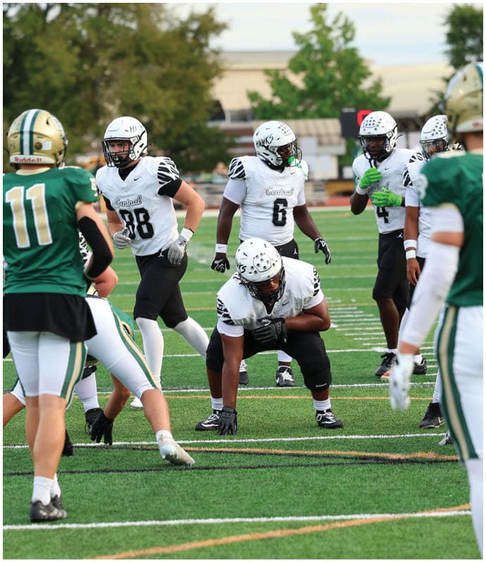 People wearing football uniforms are on a field with one person in a green jersey preparing to throw a ball while others in white jerseys are positioned defensively. Descripci n generada con IA