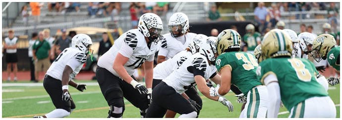 People in football uniforms are positioned on a field facing each other, preparing for a play during a game. Descripci n generada con IA