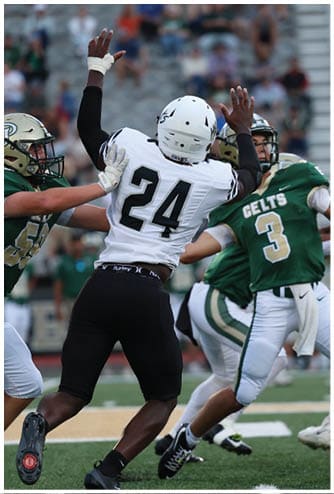 Two people in football uniforms are engaged in a play on a football field with one person in a green jersey throwing a ball and another in a white jersey attempting to block or catch it. Descripci n generada con IA