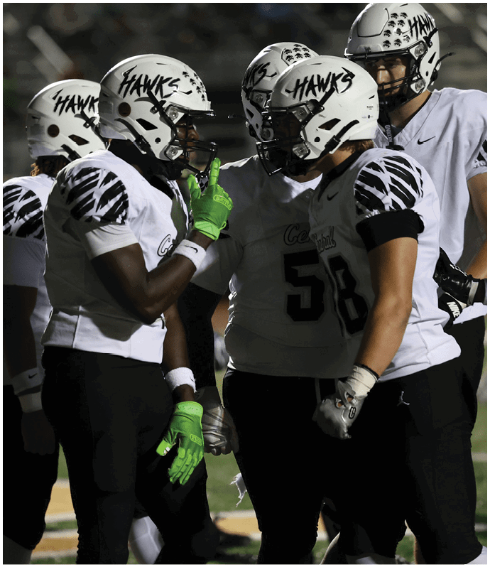Three people wearing white football helmets with “HAWKS" written on them, white jerseys with black patterns on the shoulders, black pants, and bright green gloves stand on a football field. Descripci n generada con IA