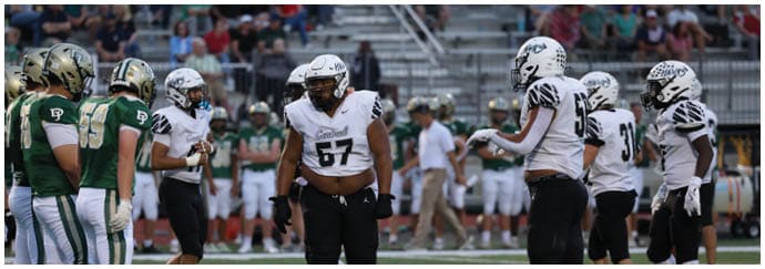 Two opposing American football teams in green and white uniforms face each other on the field with players and coaches in the background. Descripci n generada con IA