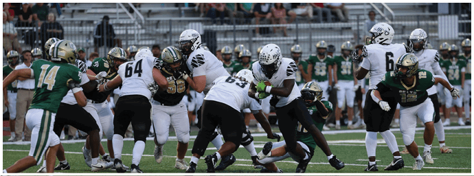 People in football uniforms are engaged in a play on a field with one player holding the ball while others block and tackle. Descripci n generada con IA