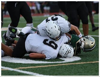 A person wearing a white football helmet with “HAWKS" written on it and a white jersey is bent over on a football field. Descripci n generada con IA