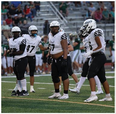 A person wearing a white football uniform with the number 57 and a helmet is standing on a field with blurred spectators in the background. Descripci n generada con IA