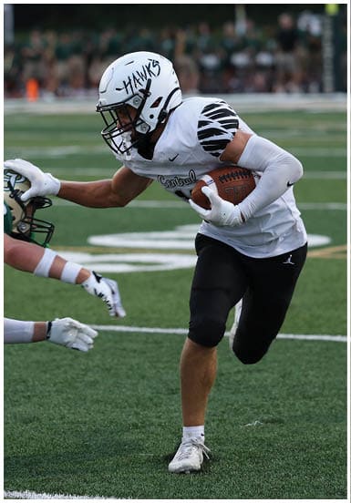 A person in a white football uniform with “HAWKS" on the helmet is running with the ball while another person in a green and white uniform reaches out to tackle them on a football field. Descripci n generada con IA