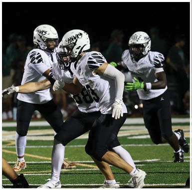 Several people wearing white football helmets and uniforms with “Hawks" written on the helmets are actively engaged in a football game on a field at night. Descripci n generada con IA