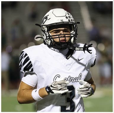 A person wearing a white football uniform and helmet with black accents is standing on a field, appearing focused and ready for action. Descripci n generada con IA
