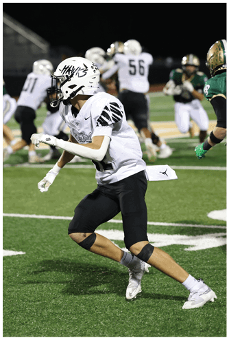 A person in a white football uniform and helmet is running on a green field during a nighttime game with other players in the background. Descripci n generada con IA